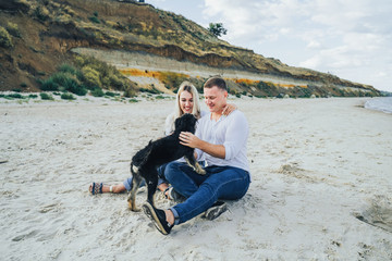 A beautiful, attractive, loving couple playing with mutts on the seashore. Lovers, wearing white coats and blue jeans, petting cheerful dogs, that ran to them. Dogs licking people and jumping high