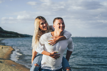 Attractive blonde girl jumped onto her boyfriend's back. He carries her, she laughs and kisses him. They run along the sand on the seashore on a summer evening, lit by the sun. Romantic Date Concept