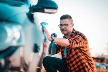 Young man polishing his car with a cloth and spray in a bottle, outdoors.