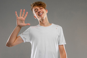 Caucasian smiling guy in a white T-shirt shows five fingers on a gray background