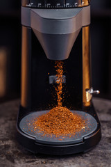 A beautiful black coffee grinder is standing on the table. Freshly grounded coffee is poured. Nearby there are two cups of espresso. A man holding out his hand for one cup of coffee