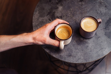 A beautiful black coffee grinder is standing on the table. Freshly grounded coffee is poured. Nearby there are two cups of espresso. A man holding out his hand for one cup of coffee