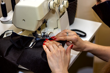 Obraz na płótnie Canvas blonde girl with a black mask on her face sews fabric protective masks on an overlock sewing machine to protect against coronavirus during an epidemic