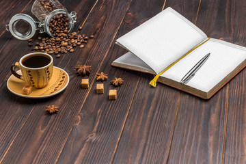 Open notebook with pen. Cup of coffee, coffee beans in glass jar. On the table spices star anise...