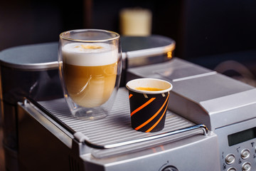 a cup of cappuccino and a cup of espresso are standing on top of an office coffee machine. Inexpensive, simple coffee machine model makes delicious coffee with milk and natural custard americano