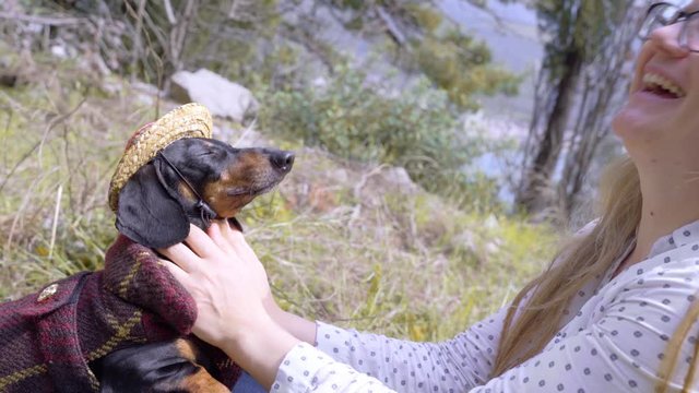 Cute dachshund in mexican costume with checkered poncho and straw wide brimmed sombrero hat. Blonde girl in glasses kisses her beloved dog and laughs on street. Mountainside and sea on the background.