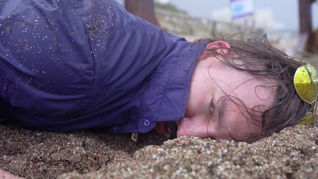 Drunk guy in blue shirt and sunglasses on his head lies face in wet sand and coughs, close up. Man saved from drowning comes to his senses, needs emergency medical care.