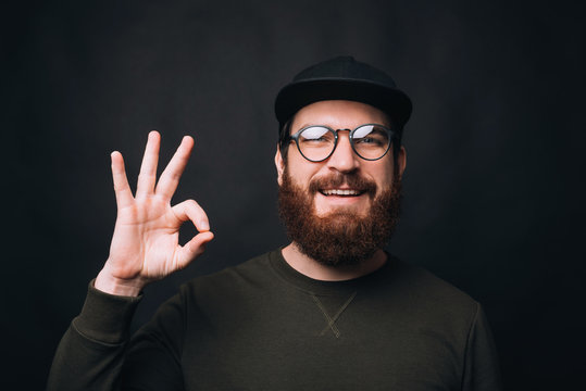 Handsome bearded man wearing glasses is showing ok gesture on black background.