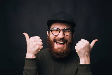 This is cool. Happy bearded man wearing glasses is showing thumbs up on black background.