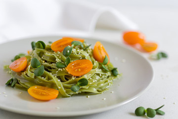 A grey plate with green pasta topped with raw orange tomatoes, sesame seeds and sunflower seeds micro greens. Horizontal orientation. 