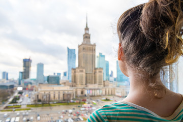 Back view of young woman standing by window looking at aerial high angle view of Warsaw, Poland cityscape downtown with Palace of Science and Culture building skycrapers