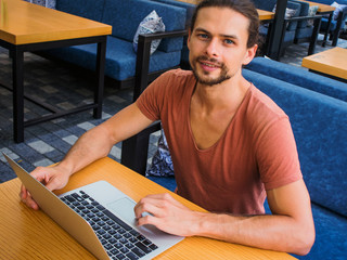 young handsome man businessman sitting in a cafe with laptop looking at you and smiling, technology online
