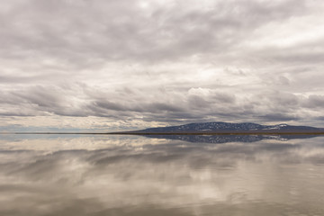 The sloping mountain on the horizon and gray clouds are reflected in the smooth surface of the water on a cloudy day