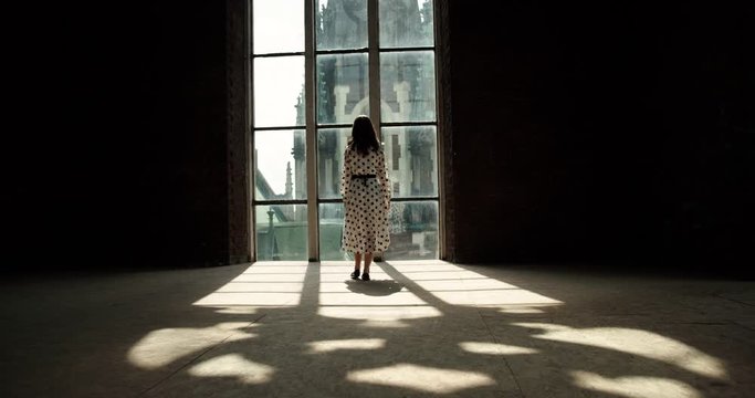 rear view. the girl slowly walks to the window. through the window into the room rays of sunlight.