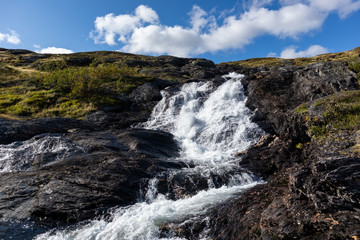 Sunny white scenic norwegian powerful river cascade waterfall with dark rocks and bright blue sky. Nature travel clean falling water landscape