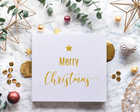 Flat lay on white marble background with text "Merry Christmas" on canvas. Christmas frame with fresh eucalyptus twigs and golden geometric decorations - hexagons, dark red trinkets and wire shapes.