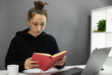 Serious beautiful young woman in black hoodie with hear in bun reading book in red cover at table...