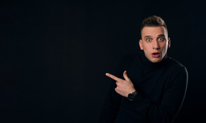 Young surprised man in black t-shirt pointing left with his finger, shouting WOW, isolated on gray background with copy space fot your text. Shock content.