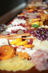 Lot of different fresh food is cut into pieces on a long tray on the table. Slicing fruits, vegetables, cheeses and sausages. Soft focus.
