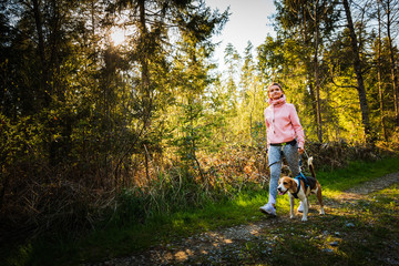 Young woman and dog walking together on country path in forest. Cheerful female exercising outdoor with her pet.