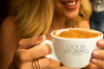 smiling young girl in a cafe  holding a big cup of hot cappuccino with coffee foam design of heart on it and words good morning on the cup