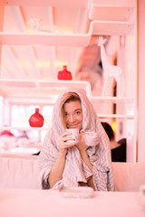 a beautiful young girl sitting in a cafe with a cover on her head as she is cold smiling and drinking hot tea trying to get warm 