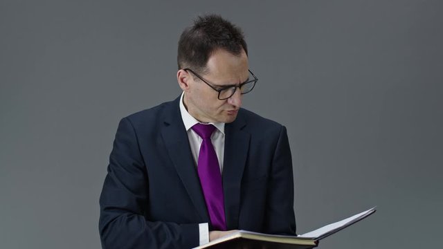 Handsome middle aged individual entrepreneur in formal clothes, tie and glasses checks his notes in daily planner and seriously looks at camera on isolated grey background, business plans concept