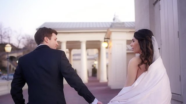 A bride and groom holding hands and walking across their reception venue brick patio in slow motion