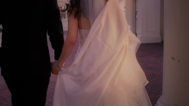 From behind a newly married couple as they walk through pillars on a brick terrace in slow motion