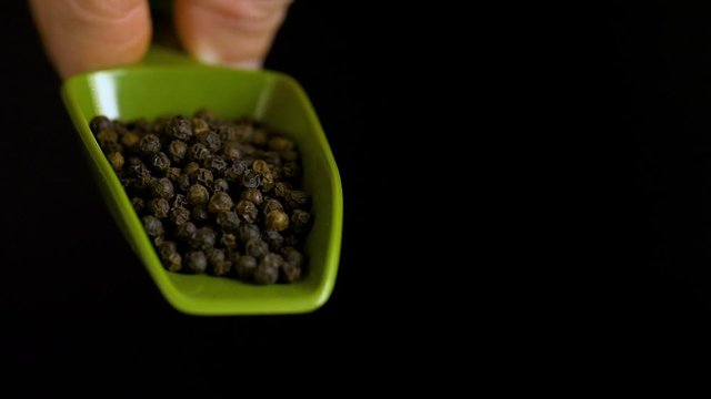 Close-up of whole black peppercorns lying on a green plastic spoon. A hand holds the spoon and presents the spice against a black background.