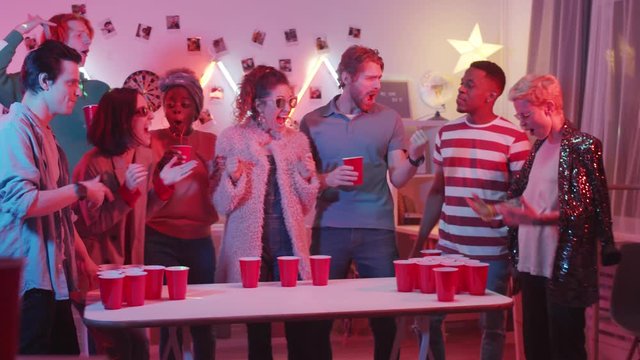 Young cheerful man and woman playing beer pong at home party with multiethnic friends. Man throwing ball into cup of female opponent and then woman drinking beer while excited friends applauding
