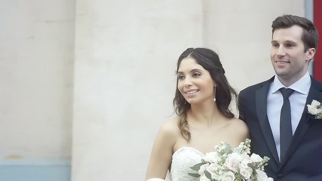 A bride holding a bouquet and a groom posing for pictures in front of a tan church wall