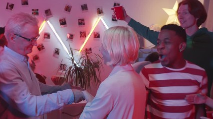 Cheerful senior couple joining young multiethnic friends and dancing at awesome house party in living room with neon light