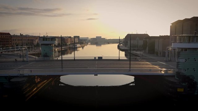 Drone video from Copenhagen harbour, flying slowly backwards from a bridge over the canal, revealing a beautiful sunrise and the city.