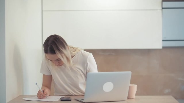 A young girl is concentrated in a laptop and outlines a white sheet of paper. Read on the calculator in the phone.