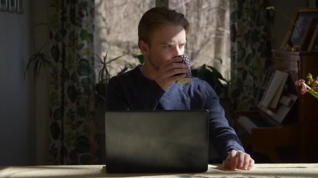 Close up portrait: young Caucasian man sits at computer, drinks coffee and thinks