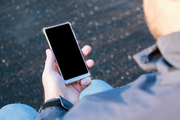 mockup sitting man's hand holding a white mobile phone with a blank screen outdoors