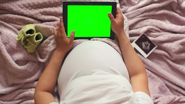 Top view of a pregnant woman lying in bed with a tablet with a green screen. Pregnant girl in bed with baby socks and ultrasound photos