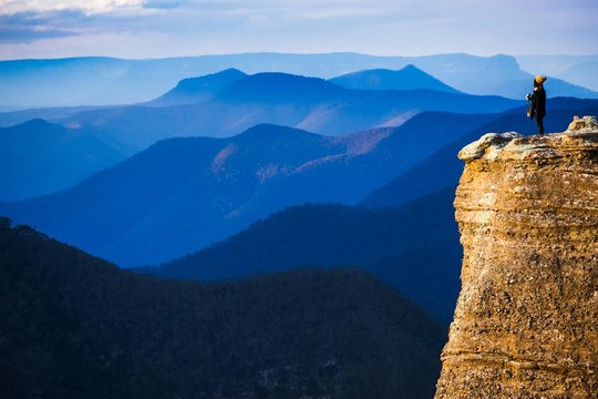 Person Standing On Rock Formation Against Blue Mountains