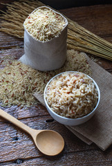 Cook and raw brown rice in white ceramic bowl on wood table and wood background with spoon. Copy space.