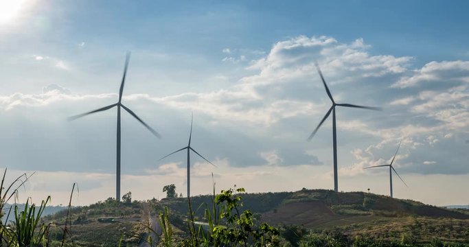 Time lapse Wind turbines on hills landscape with green meadow, hill with flowers, blue cloudy sky with mist.