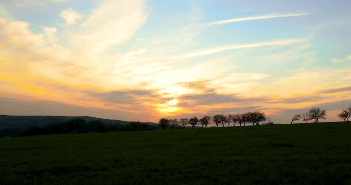 The deciduous tree line of an apple tree during a dark sunset with many colors when the sun shines through the clouds to the field and the surrounding grass through the steam line to compete 4k 60fps
