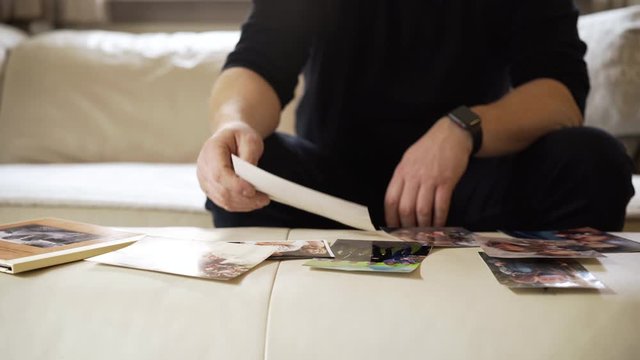 Unrecognizable male person sits on a couch in the living room and looks through old photos and related memories. Panning shot.