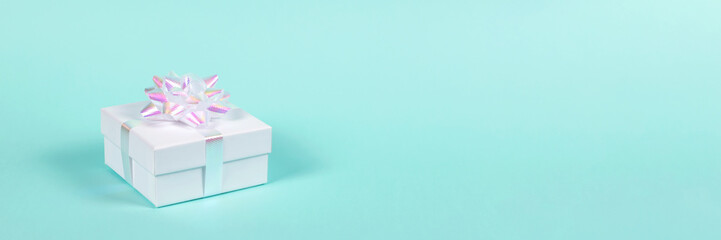 White gift box with festive holographic bow on pastel blue background. Banner for website.