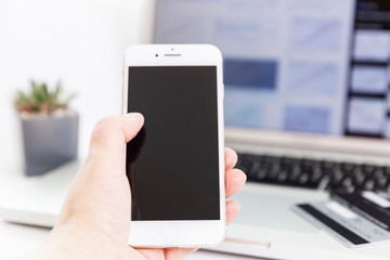 A person is holding a smartphone in their hand in front of a laptop with graphs open in it and a potted plant nearby. Perfect for business.
