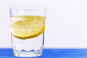 A tall glass of transparent water with a lemon slice in it on a white background with a blue stripe at the bottom.