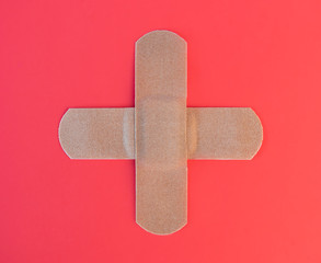 Real fabric bandages in shape of a medical plus sign. Symbol for: Hospital, emergency, first aid, medical or health. The adhesive cloth plaster are on top of a red gradient background. Photograph.