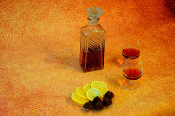 A crystal bottle and two glasses with cognac on a brown background, next to a plate with chocolates...