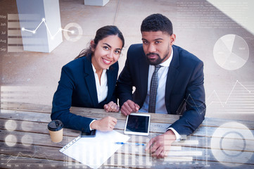 People with tablet, document and virtual statistic graphics. Man and woman wearing formal clothes, looking at camera and sitting at office desk. Business work concept. High angle view front view.