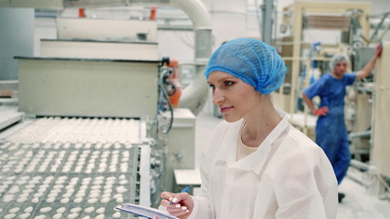 Candy factory. Controller checking conveyor with candies. Young woman in uniform holding folder and inspecting conveyor belt with fresh candies in confectionery factory.